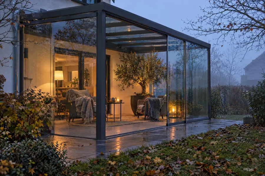 Kaltwintergarten mit Glasschiebewänden an einem nebligen Herbstabend, innen warmes Licht.