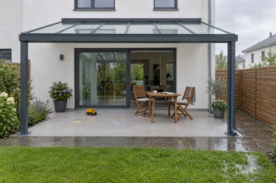 Glasdach an einem Reihenhaus bei Regen, darunter eine trockene Terrasse mit Kreidebildern.